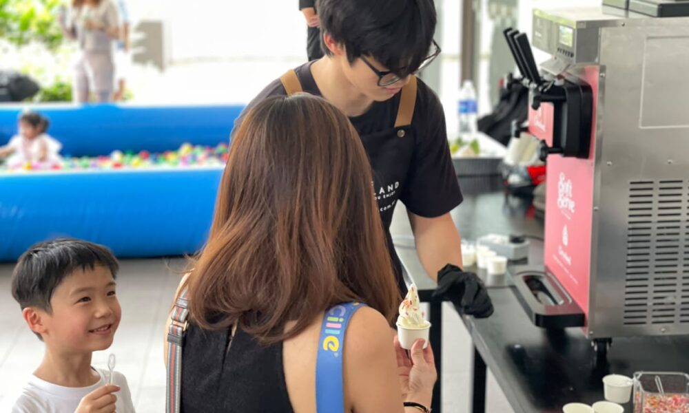 Crew member in black uniform serving soft serve ice cream to customers at an event with blue ball pit and carnival games in background