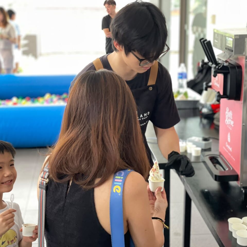 Crew member in black uniform serving soft serve ice cream to customers at an event with blue ball pit and carnival games in background